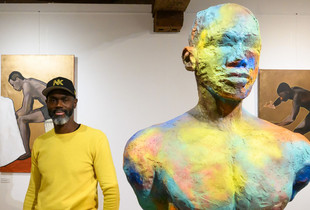 L'artiste Nald Kib devant deux tableaux et à côté d'une sculpture de son exposition