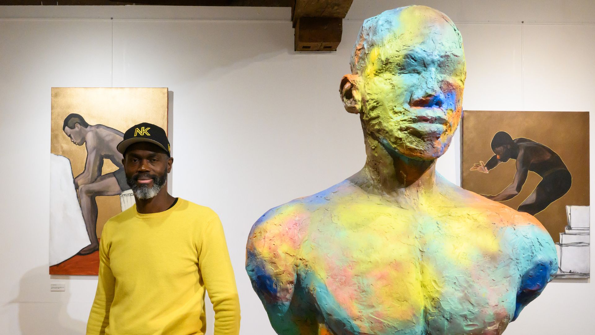 L'artiste Nald Kib devant deux tableaux et à côté d'une sculpture de son exposition